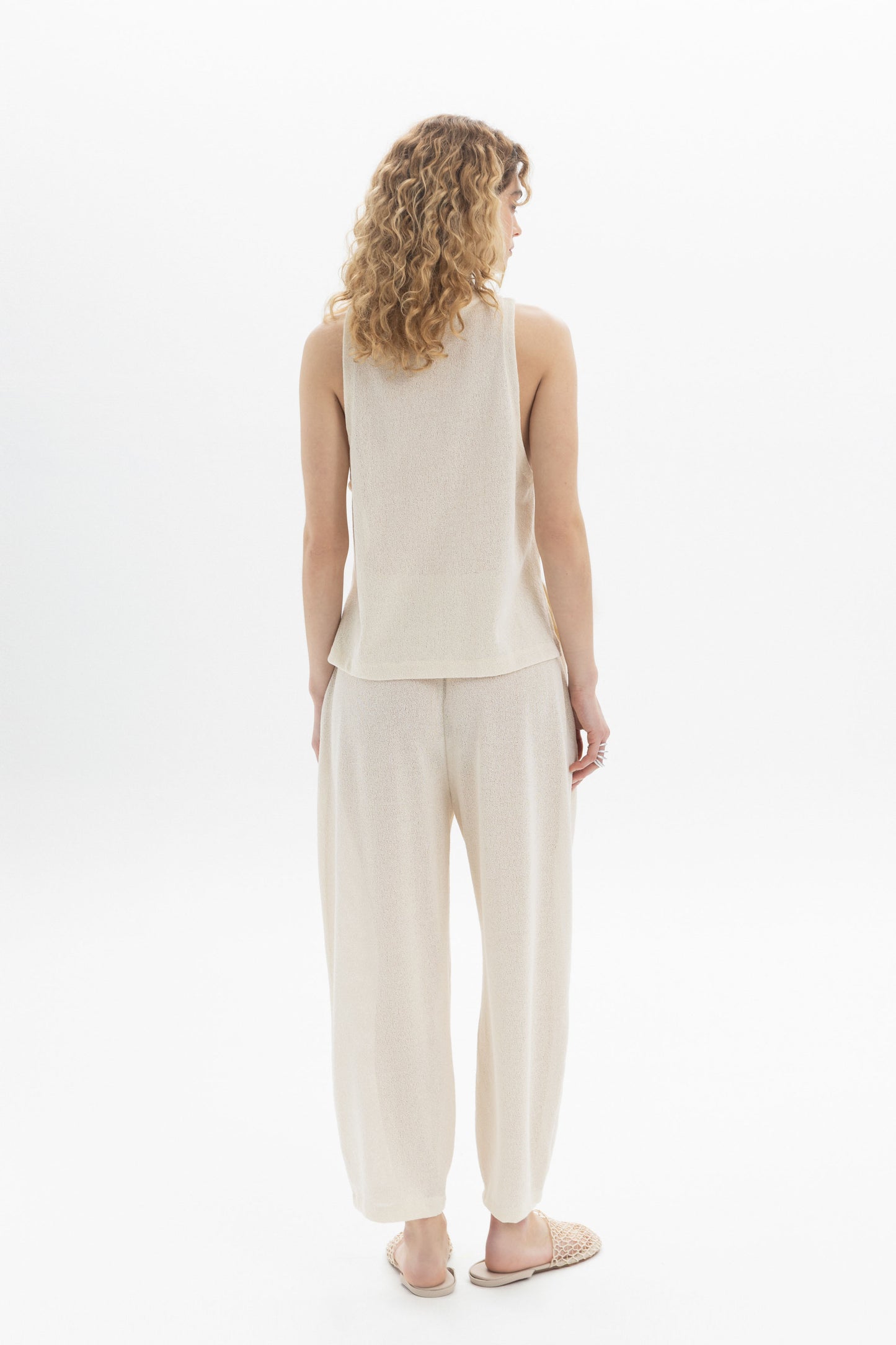 Woman seen from behind wearing a sleeveless textured top and relaxed pants in a light color.