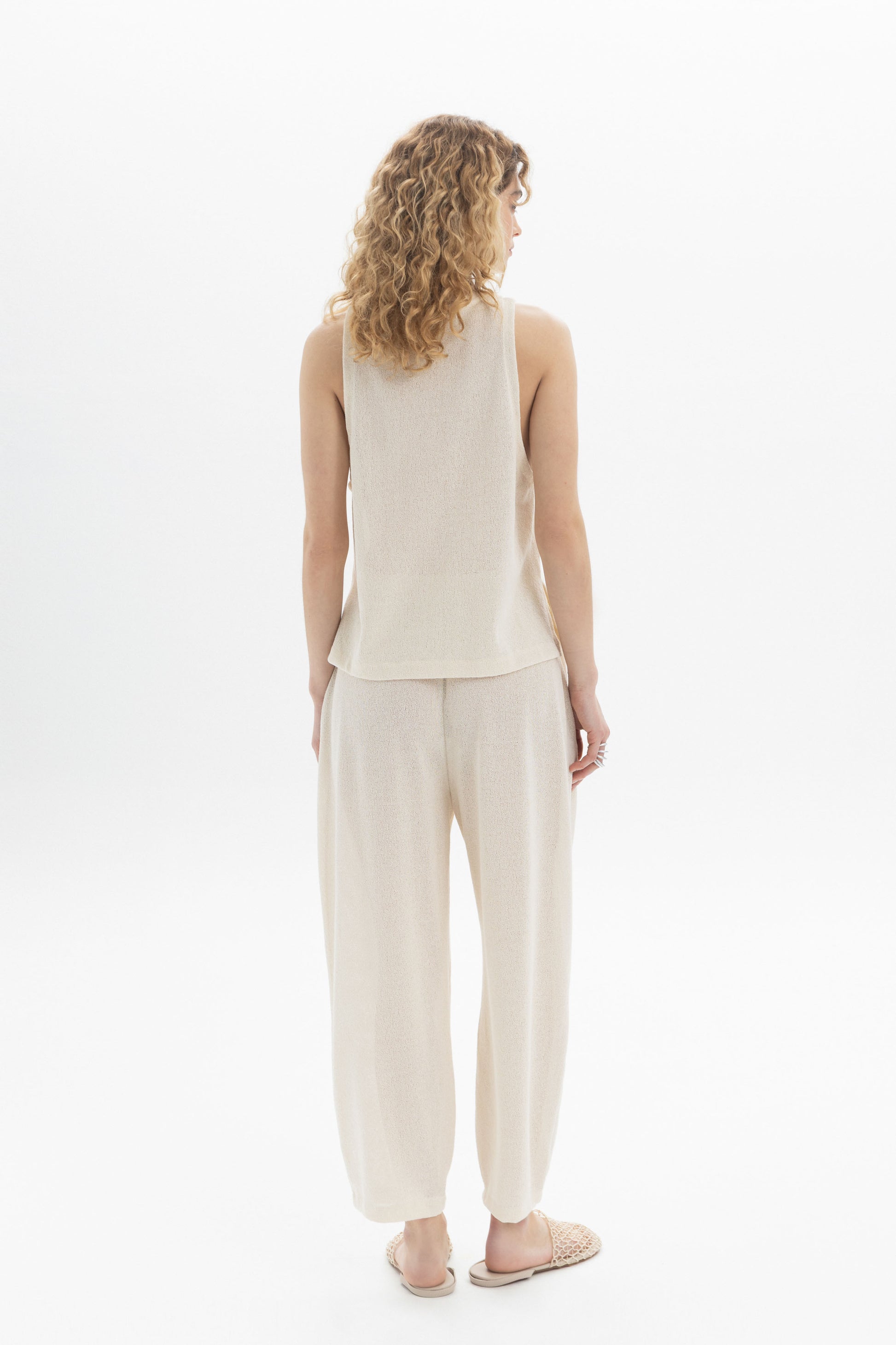 Woman seen from behind wearing a sleeveless textured top and relaxed pants in a light color.