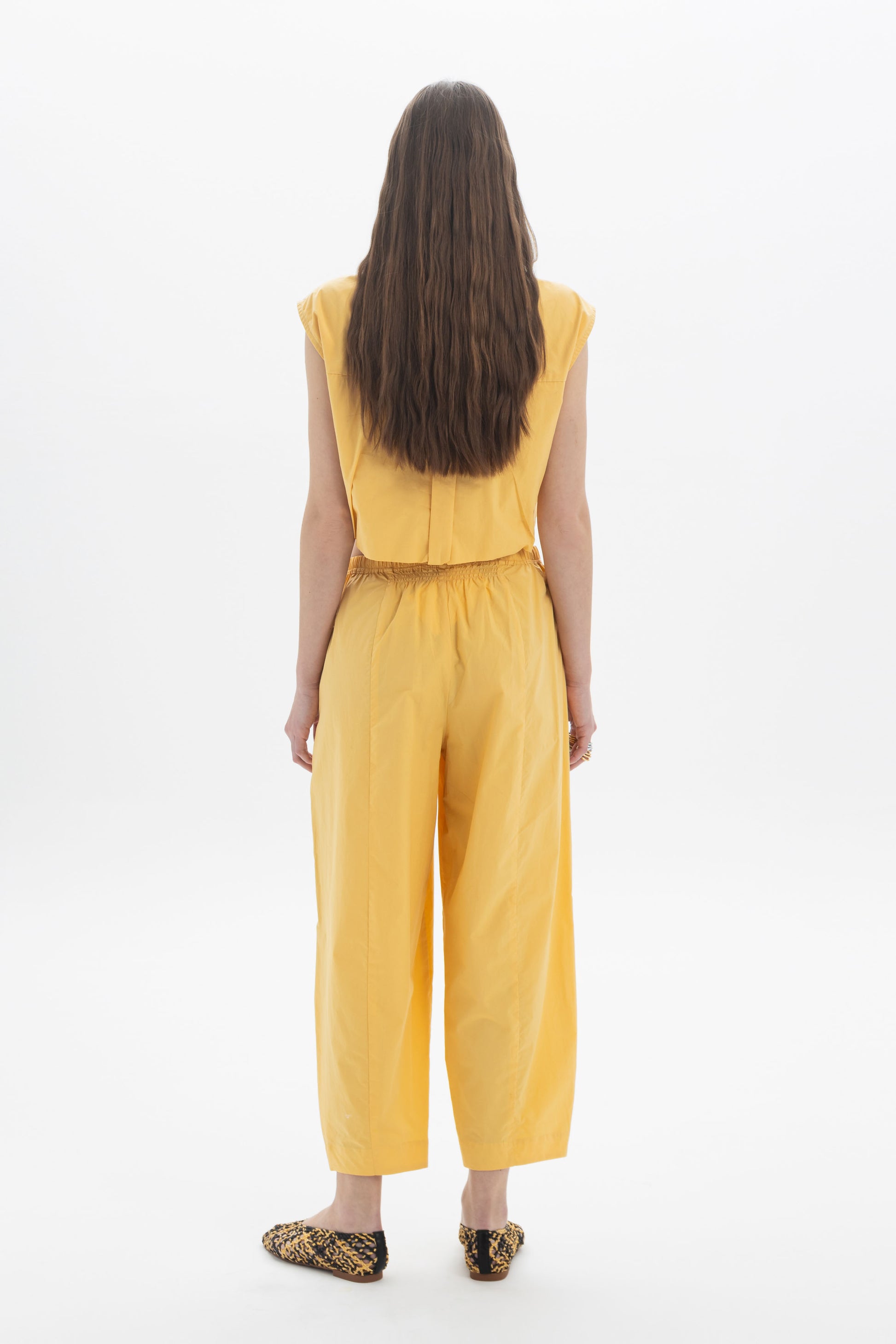 Back view of a woman wearing a yellow cropped sleeveless shirt and matching trousers, showcasing a modern outfit.