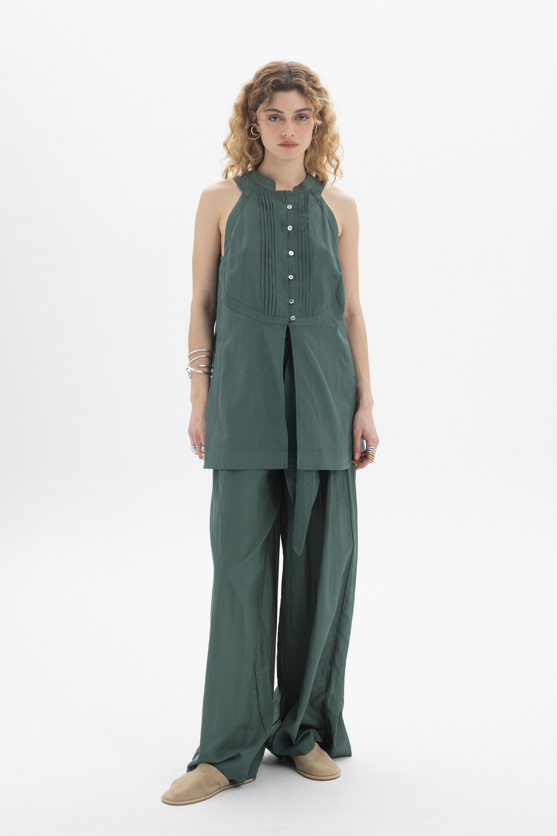 Model wearing a Sleeveless Bib Shirt in green cotton with pleat detailing and button closure, styled with wide-leg pants.
