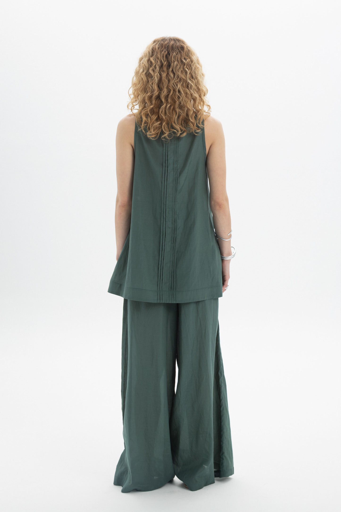 Back view of a woman wearing a green sleeveless bib shirt with a relaxed silhouette and pleat detailing.